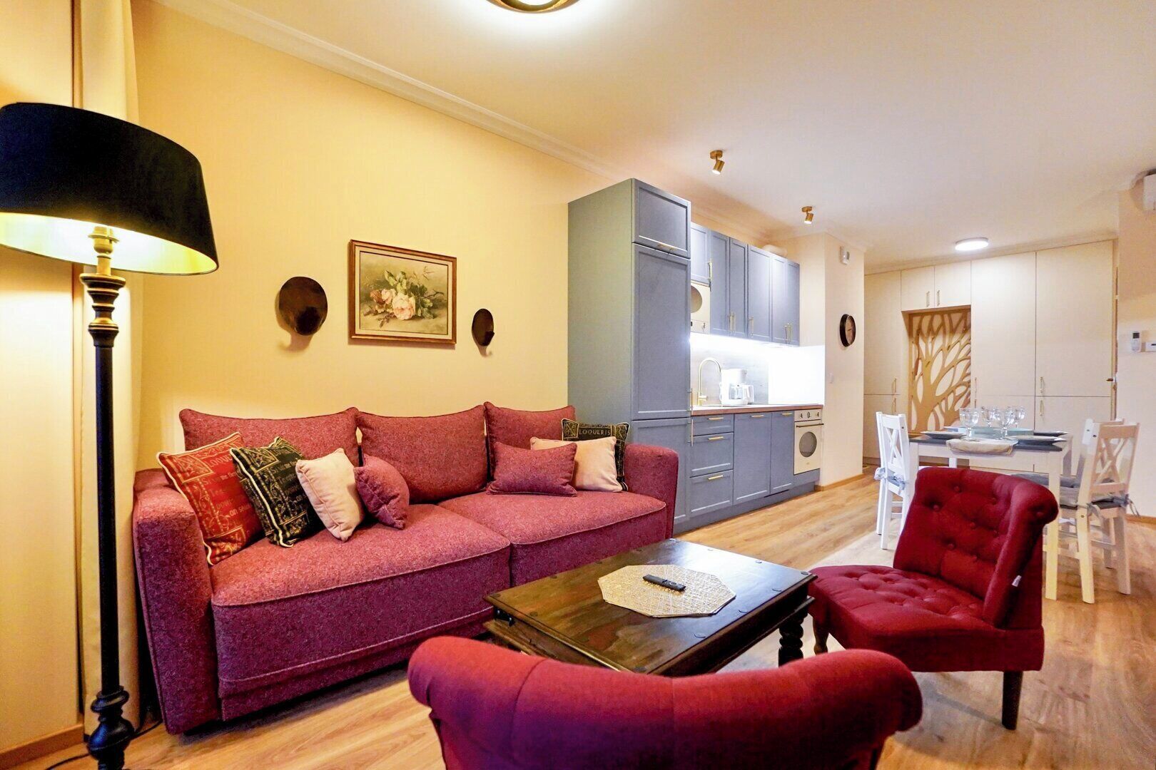 A living room with a sofa, a red armchair, a coffee table, and a lamp. There is a kitchen area with cabinets and a dining table with chairs.