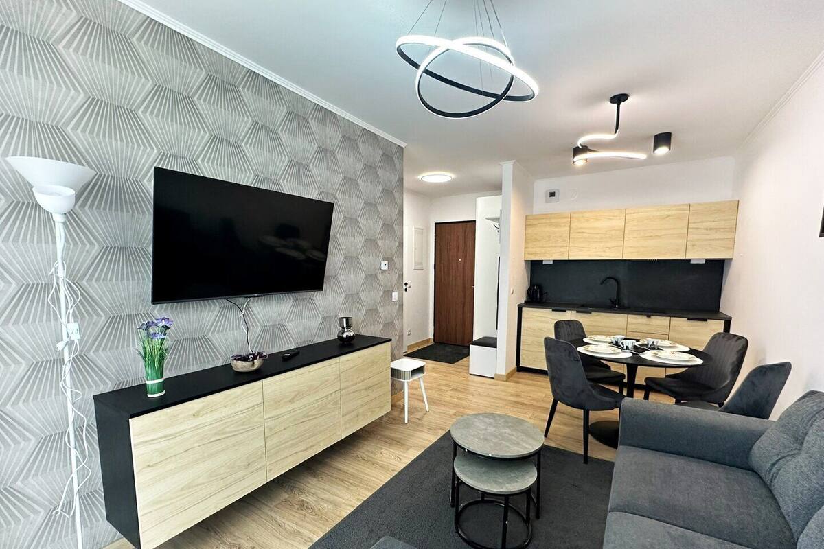 A modern living room with a flat-screen TV, a dining area with a round table and chairs, a kitchen with wooden cabinets, and a grey sofa.