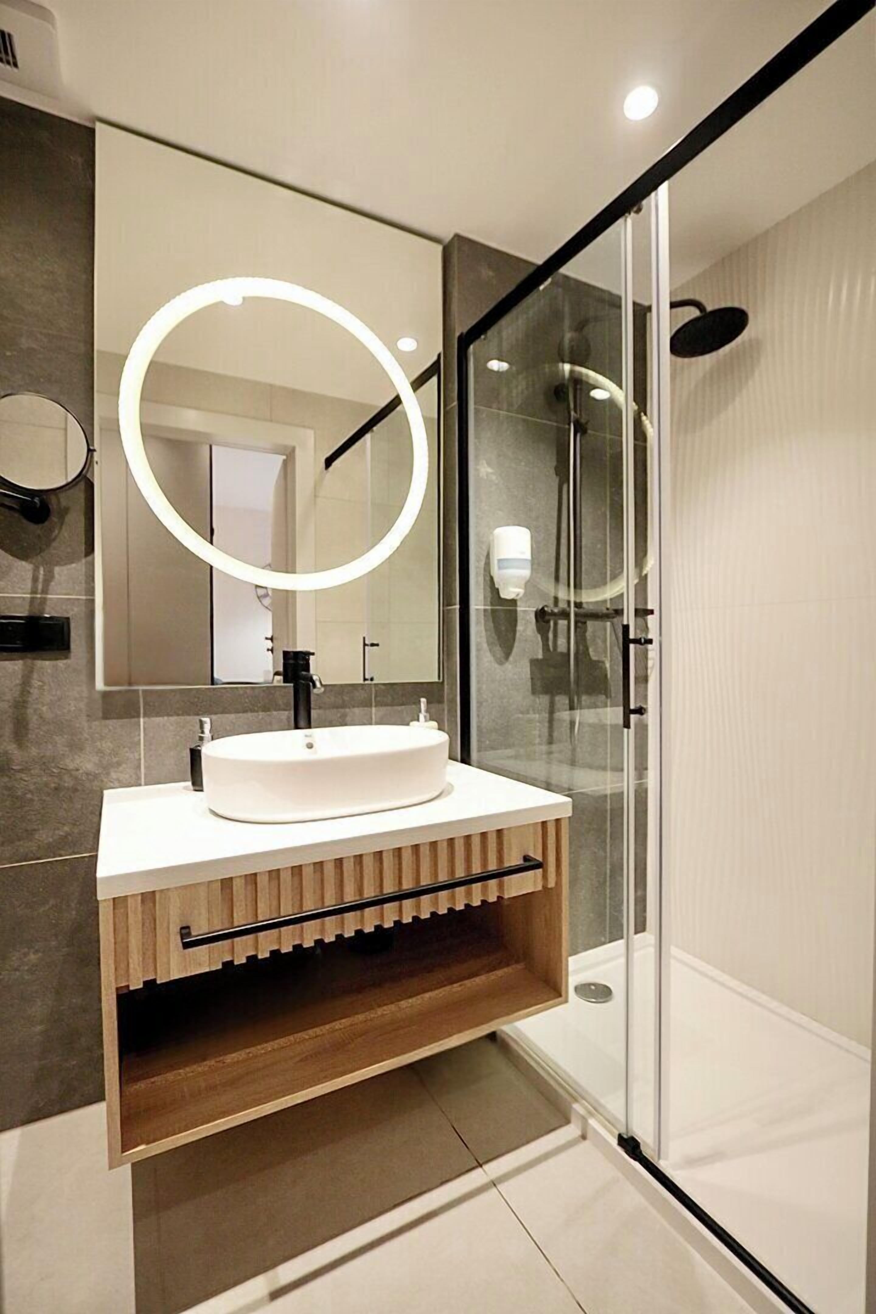 Un bagno moderno con doccia vetrata, lavandino bianco con specchio rotondo e mobile contenitore in legno.