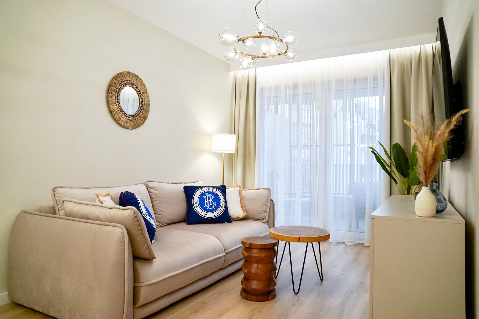 A living room with a beige sofa, a round wooden side table, a mirror, and a vase with pampas grass.