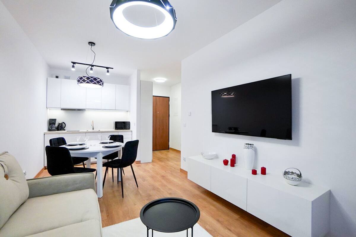 A modern living room with a sofa, a dining area with a table and chairs, a kitchen with white cabinets, a TV mounted on the wall, and a ceiling light fixture.