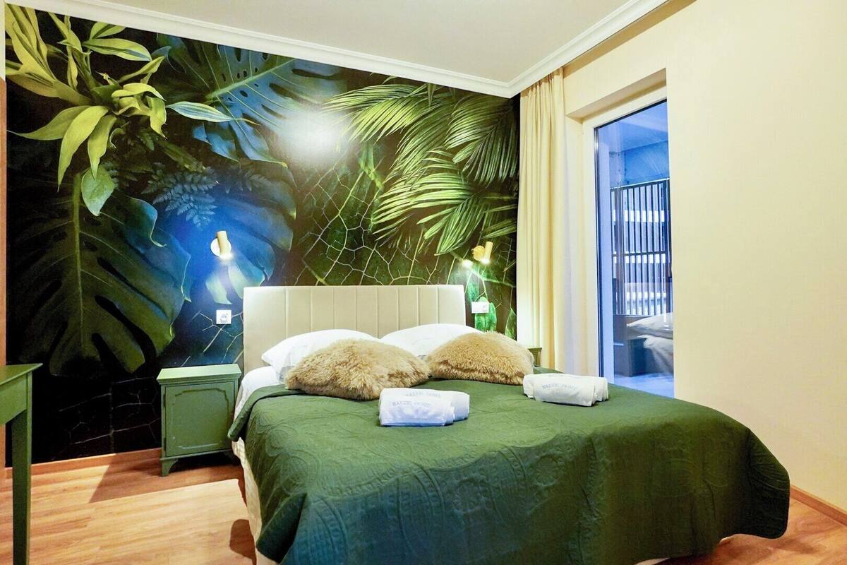 A hotel room with a bed, green bedspread, and a mural of tropical leaves on the wall.