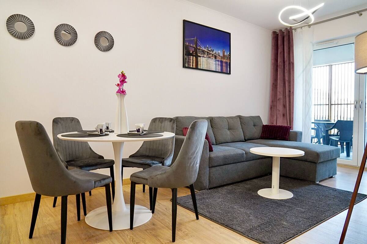 A modern living room with a grey sofa, two round tables, and matching chairs. There is a cityscape painting on the wall and a vase with flowers on a side table.