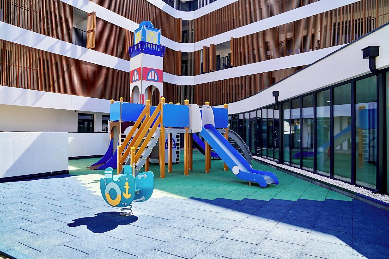 A children's play area with slides and climbing structures, surrounded by glass walls and wooden paneling.
