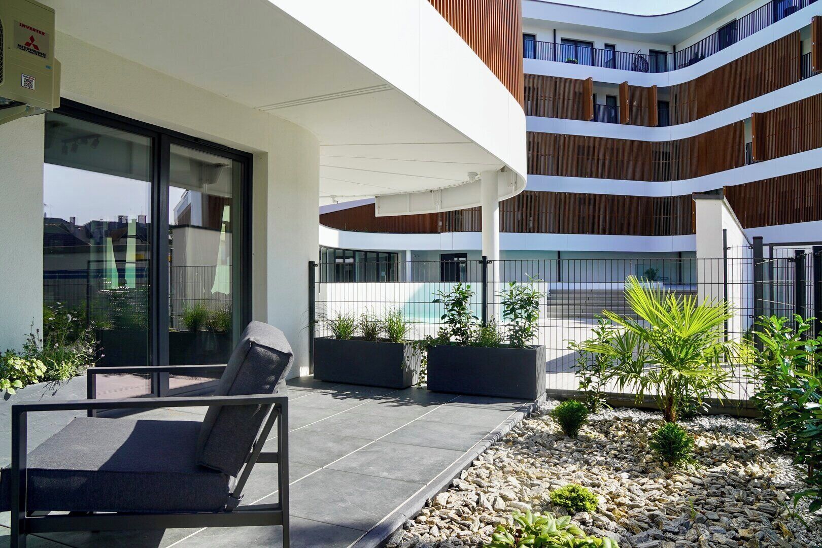 A modern outdoor area with a seating arrangement, potted plants, and a view of a multi-story building.