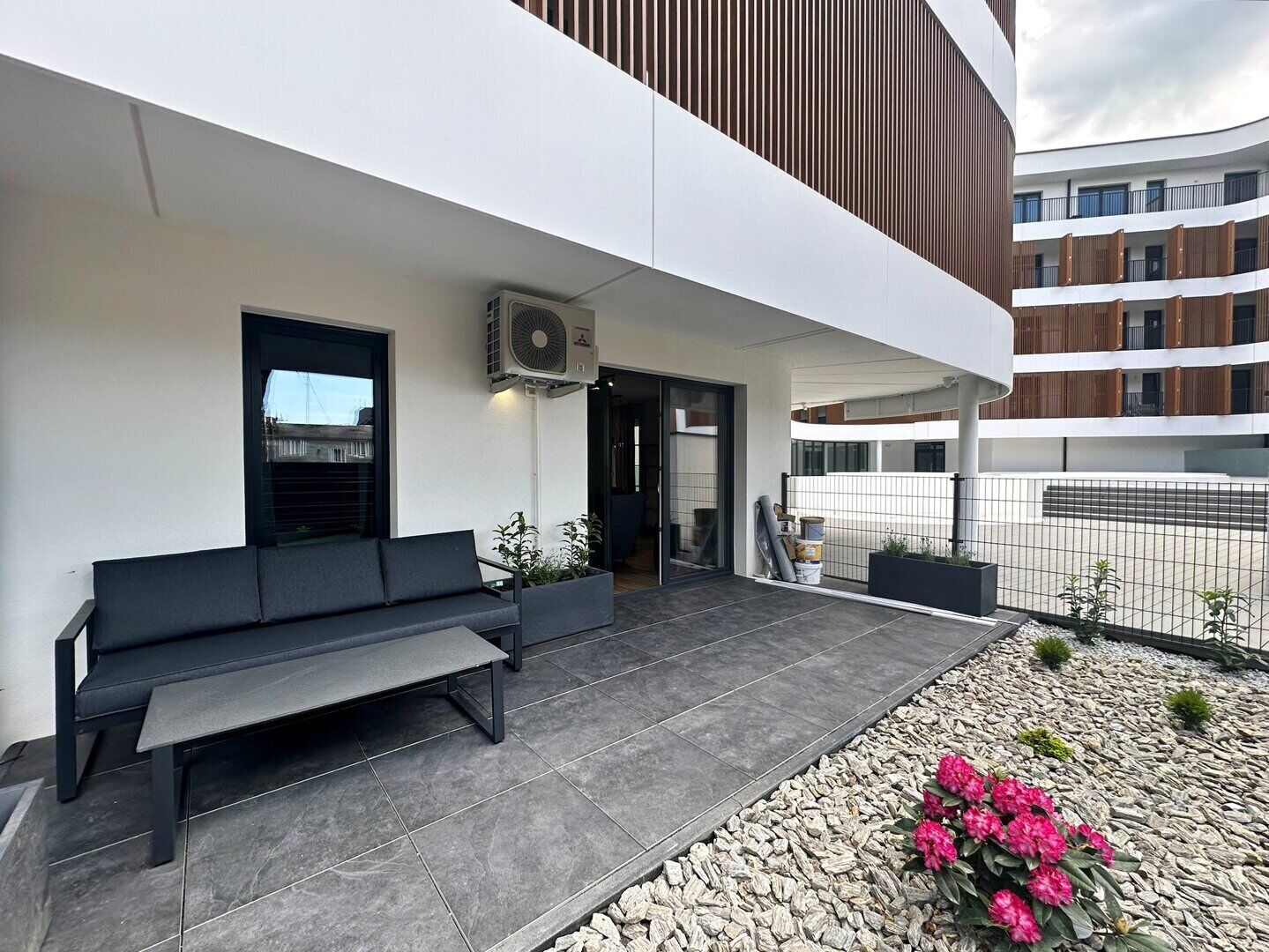 A modern building with a flat roof, a black outdoor seating area, and a small garden with pink flowers.