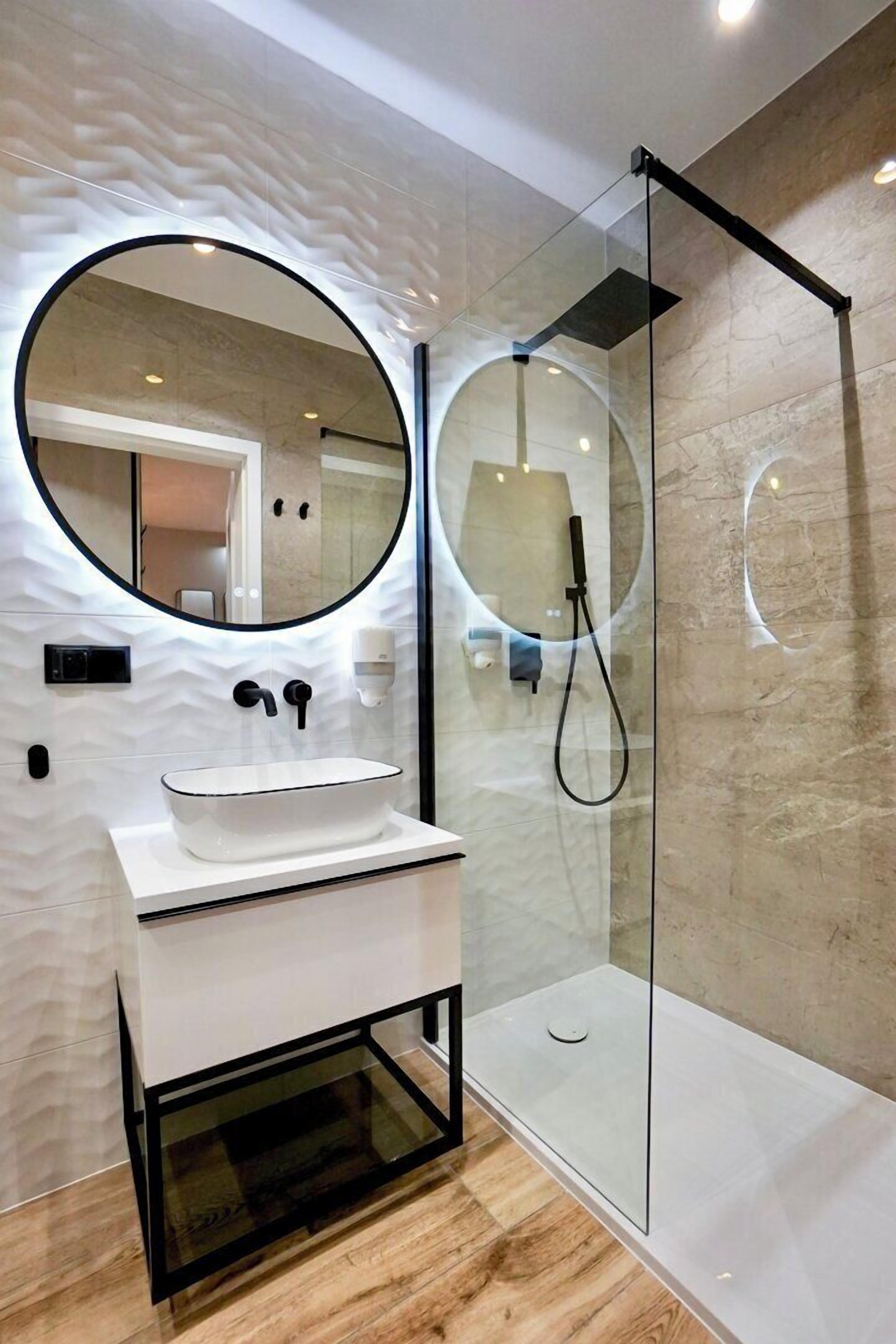 A modern bathroom with a glass-enclosed shower, a round mirror, and a white sink.