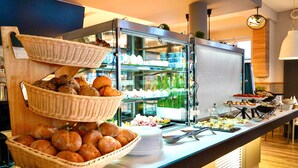 Monipuolinen buffet, jossa on tarjolla erilaisia ruokia, kuten leipää, leivonnaisia, sekä lasivitriini, jossa on vielä lisää vaihtoehtoja.