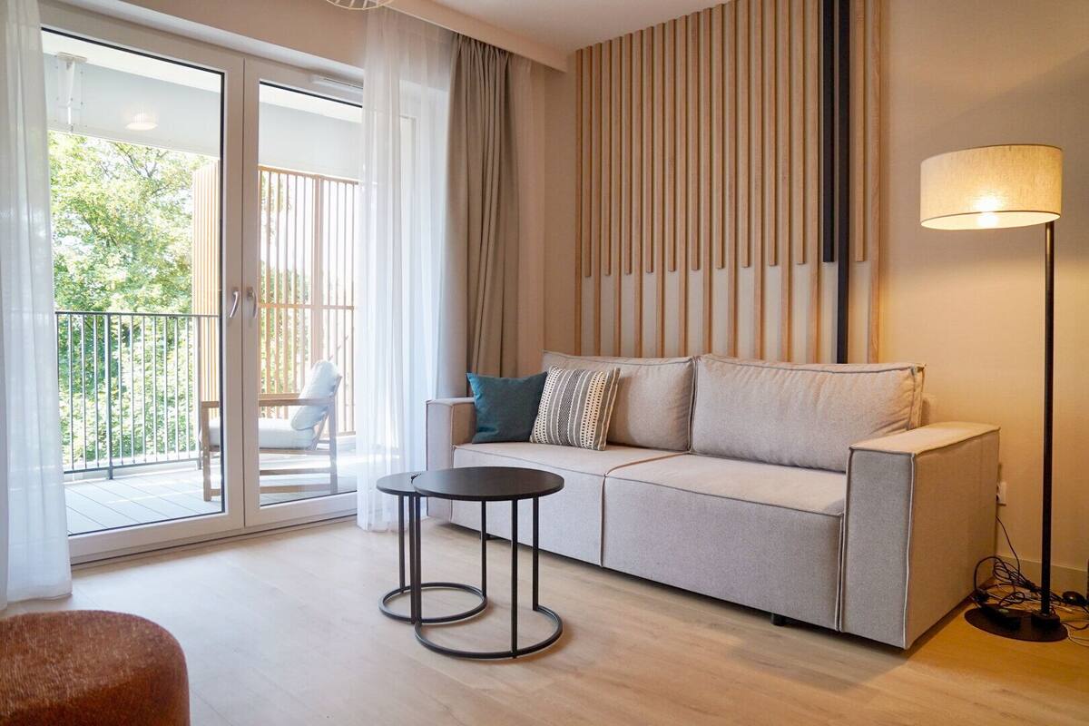 A modern living room with a sofa, a small round table, and a lamp. There is a balcony with a chair and a view of greenery outside.