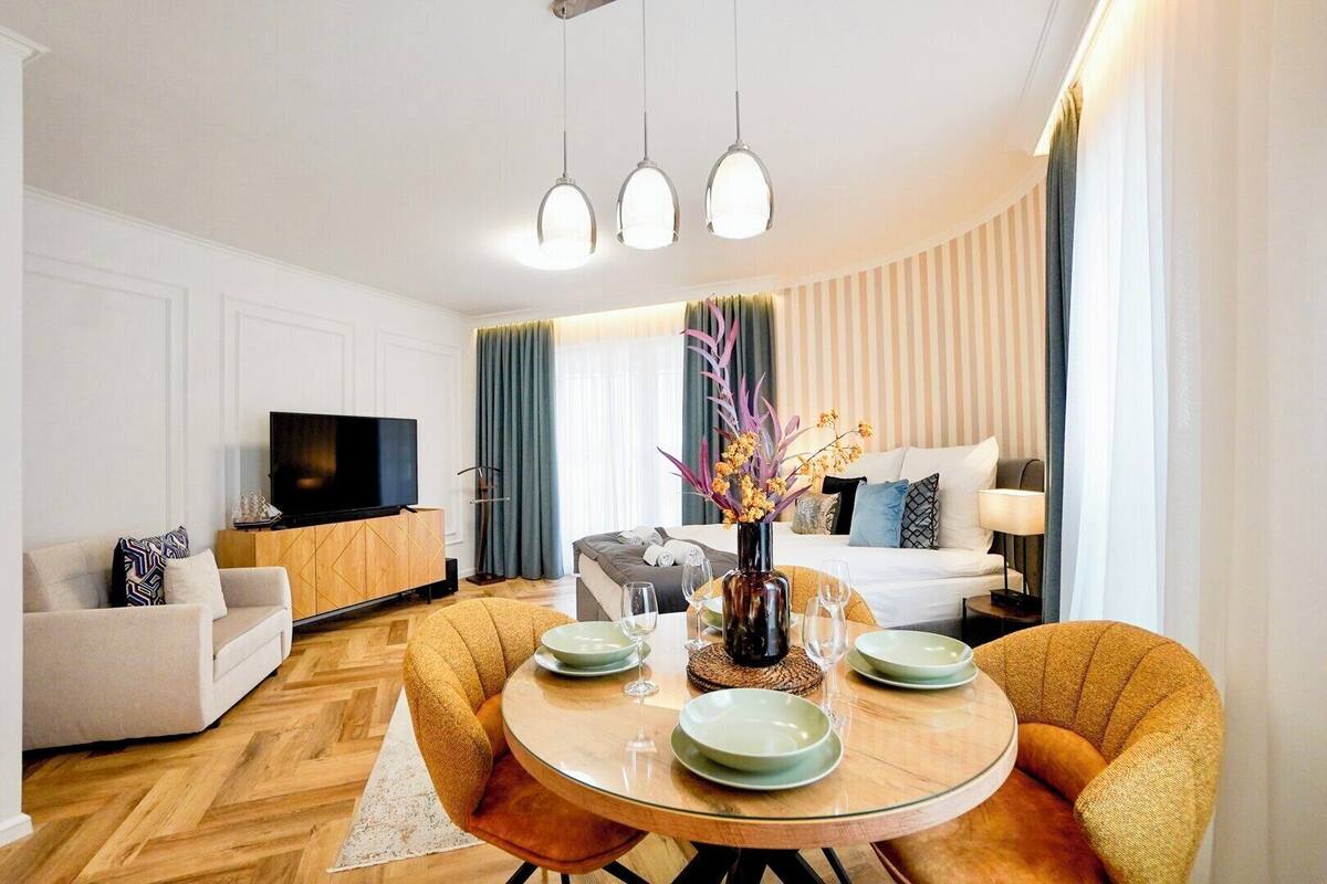 A modern hotel room with a sofa, a dining table with plates and glasses, a TV, and a bed with striped curtains.