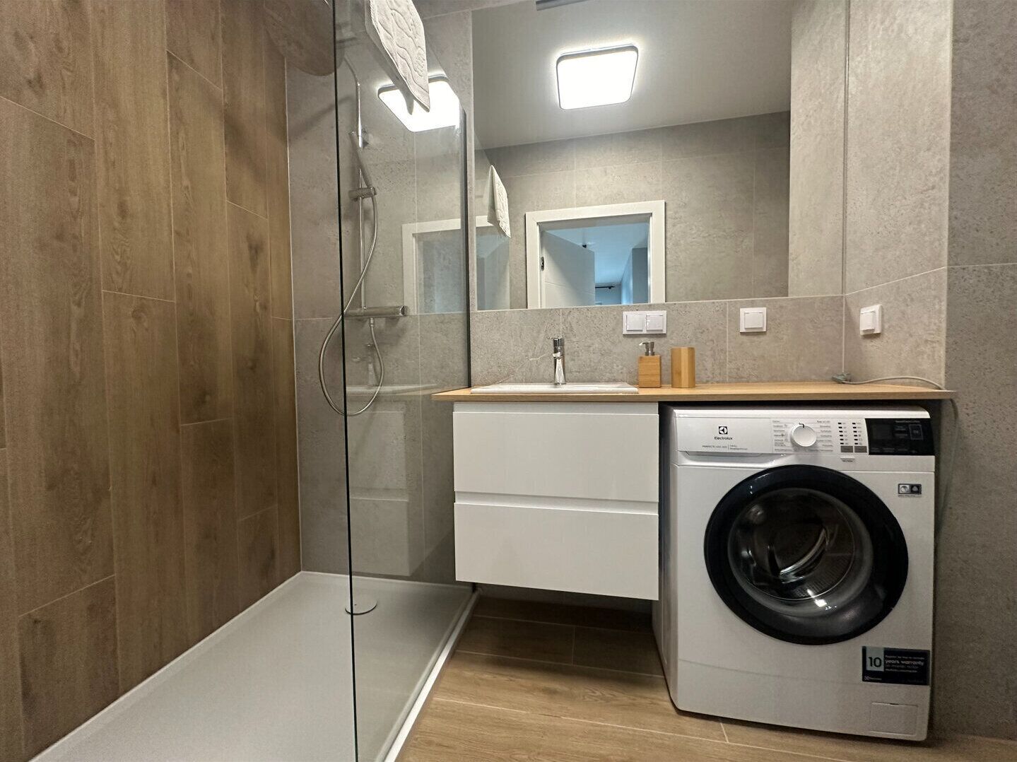 A modern bathroom with a glass shower enclosure, a built-in washing machine, and a wooden vanity.