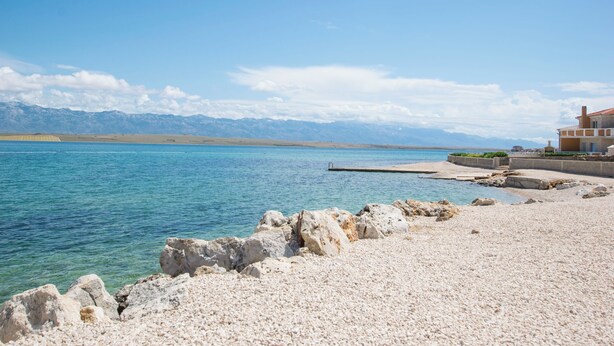 A coastal scene with a pebbly beach, a rocky breakwater, and a building with a red roof. - Villa Sana, on the beach with swimming pool (Vir)