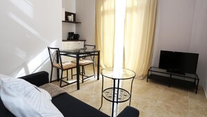 Un espai vital compacte amb un sofà, una taula de dinar amb tapa de vidre, una televisió sobre un suport i una zona de cuina amb una màquina de cafè.