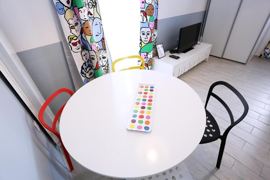 A round table with a colorful dot grid in the center, surrounded by chairs, a television on a stand, and a window with curtains featuring cartoon characters.