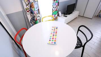 A round table with a colorful dot grid in the center, surrounded by chairs, a television on a stand, and a window with curtains featuring cartoon characters.