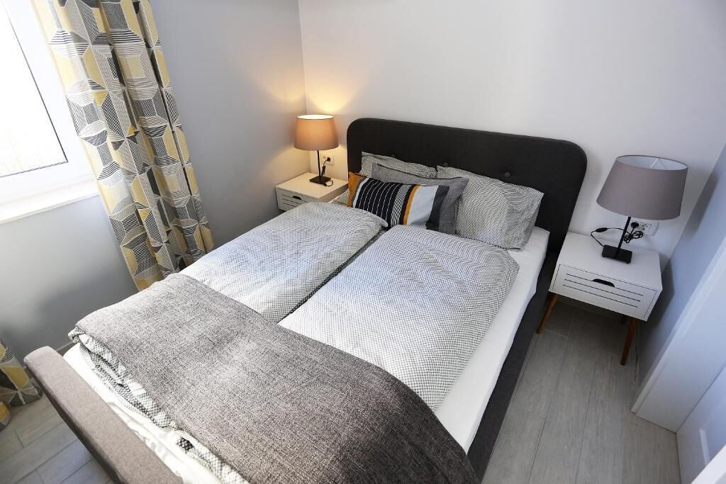 A neatly made double bed with a grey and white patterned bedspread, two pillows, a bedside table with a lamp, and a curtain with a geometric pattern.