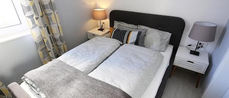 A neatly made double bed with a grey and white patterned bedspread, two pillows, a bedside table with a lamp, and a curtain with a geometric pattern.