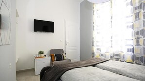 Un dormitori modern amb llit, taula de nit amb làmpada, televisió penjada a la paret i finestra amb cortines estampades.