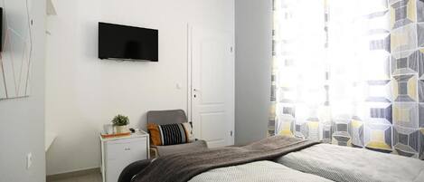 A modern bedroom with a bed, a bedside table with a lamp, a TV mounted on the wall, and a window with patterned curtains.