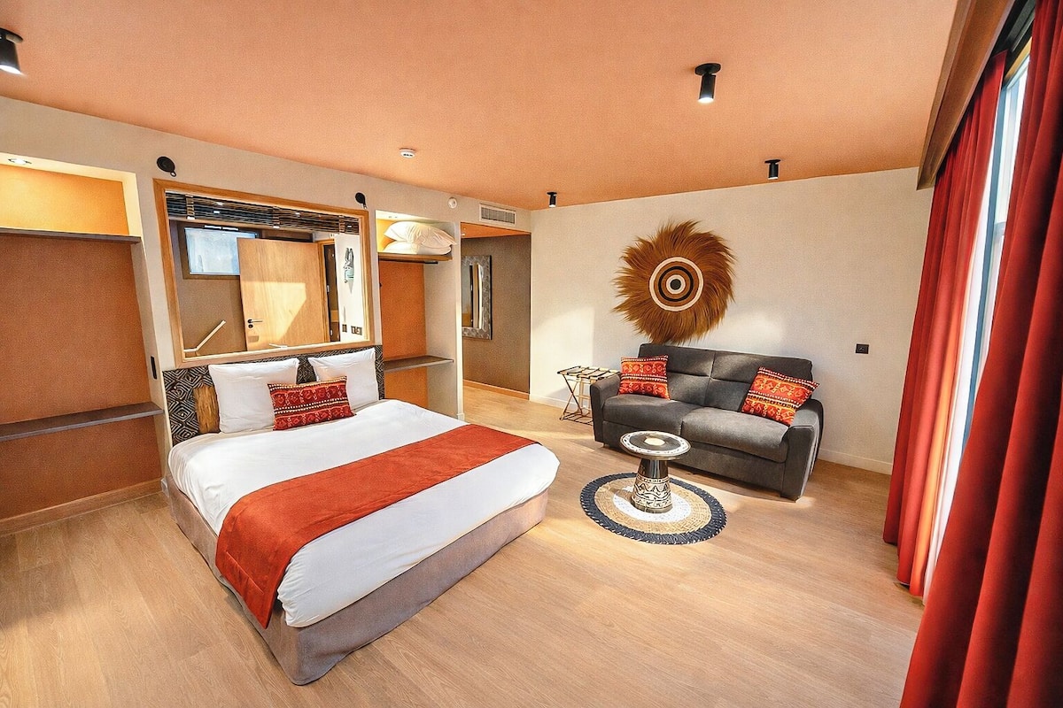 A modern hotel room with a bed, a sofa, a small table, and a decorative wall piece.