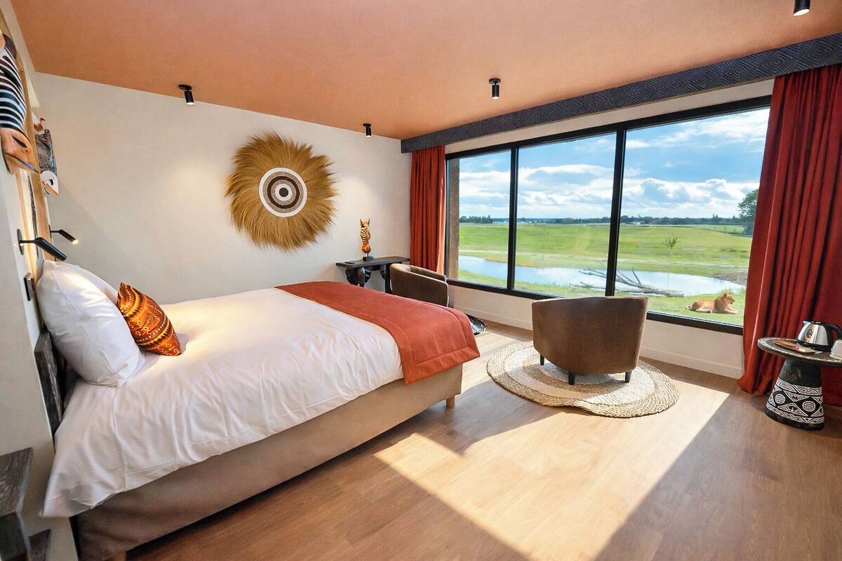 A modern hotel room with a large bed, a view of a green landscape, a round decorative wall piece, and a small table with a kettle.