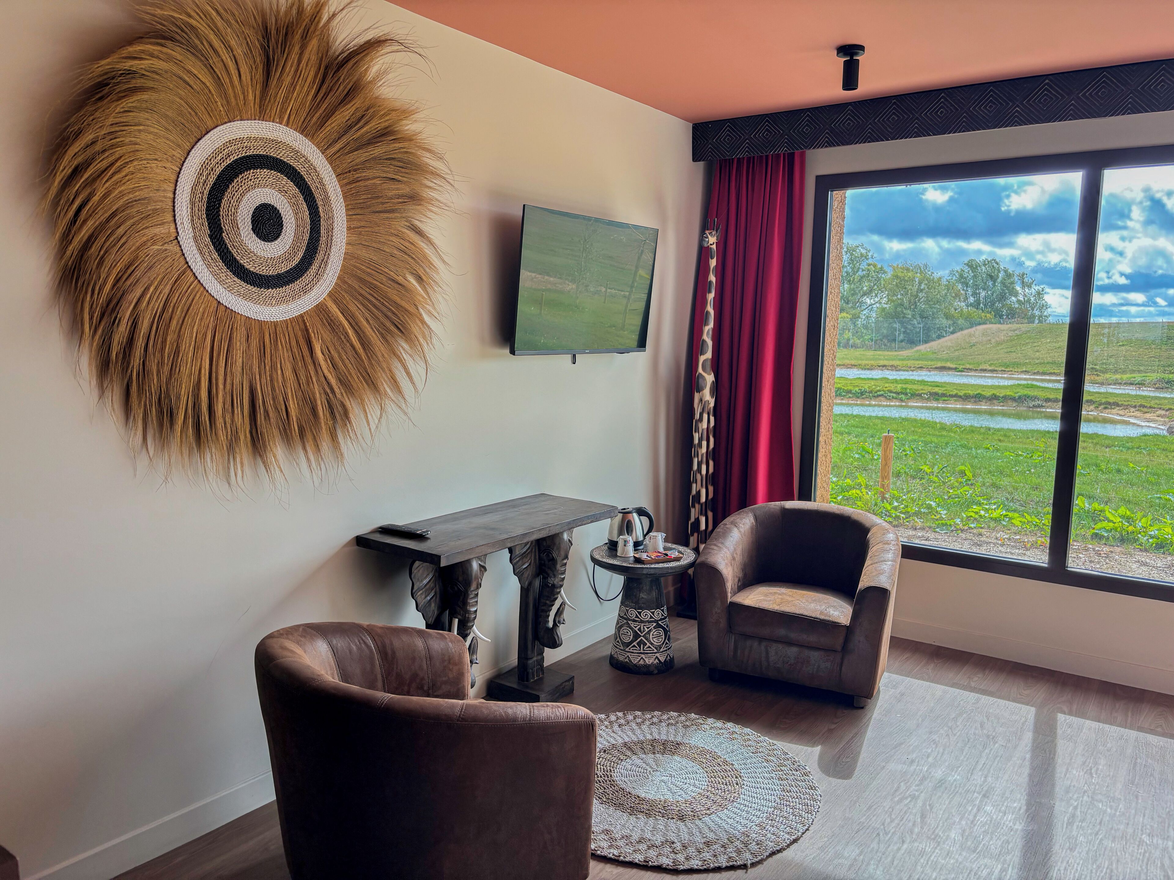 A modern living room with a large window, a mounted television, a decorative wall piece, two leather armchairs, a coffee table, and a small side table with a kettle and cups.