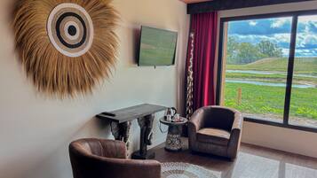 A modern living room with a large window, a mounted television, a decorative wall piece, two leather armchairs, a coffee table, and a small side table with a kettle and cups.