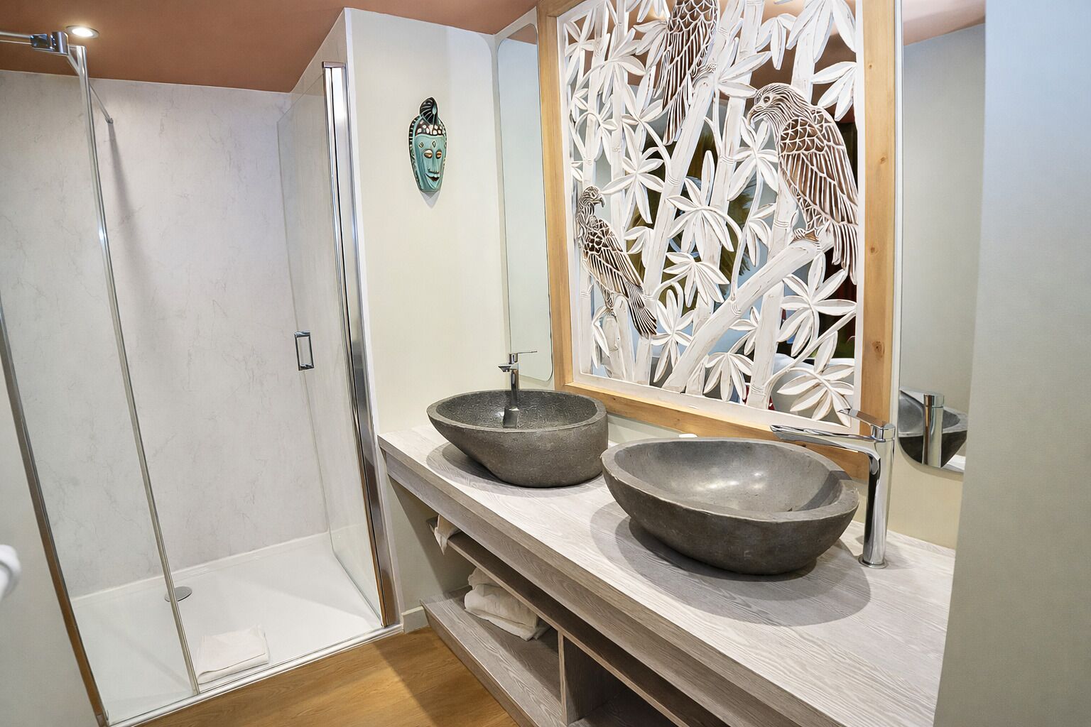 Un baño con ducha, dos lavamanos sobre encimera, un mueble de tocador de madera y un panel decorativo con pájaros.