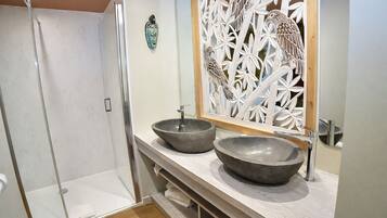Un baño con ducha, dos lavamanos sobre encimera, un mueble de tocador de madera y un panel decorativo con pájaros.