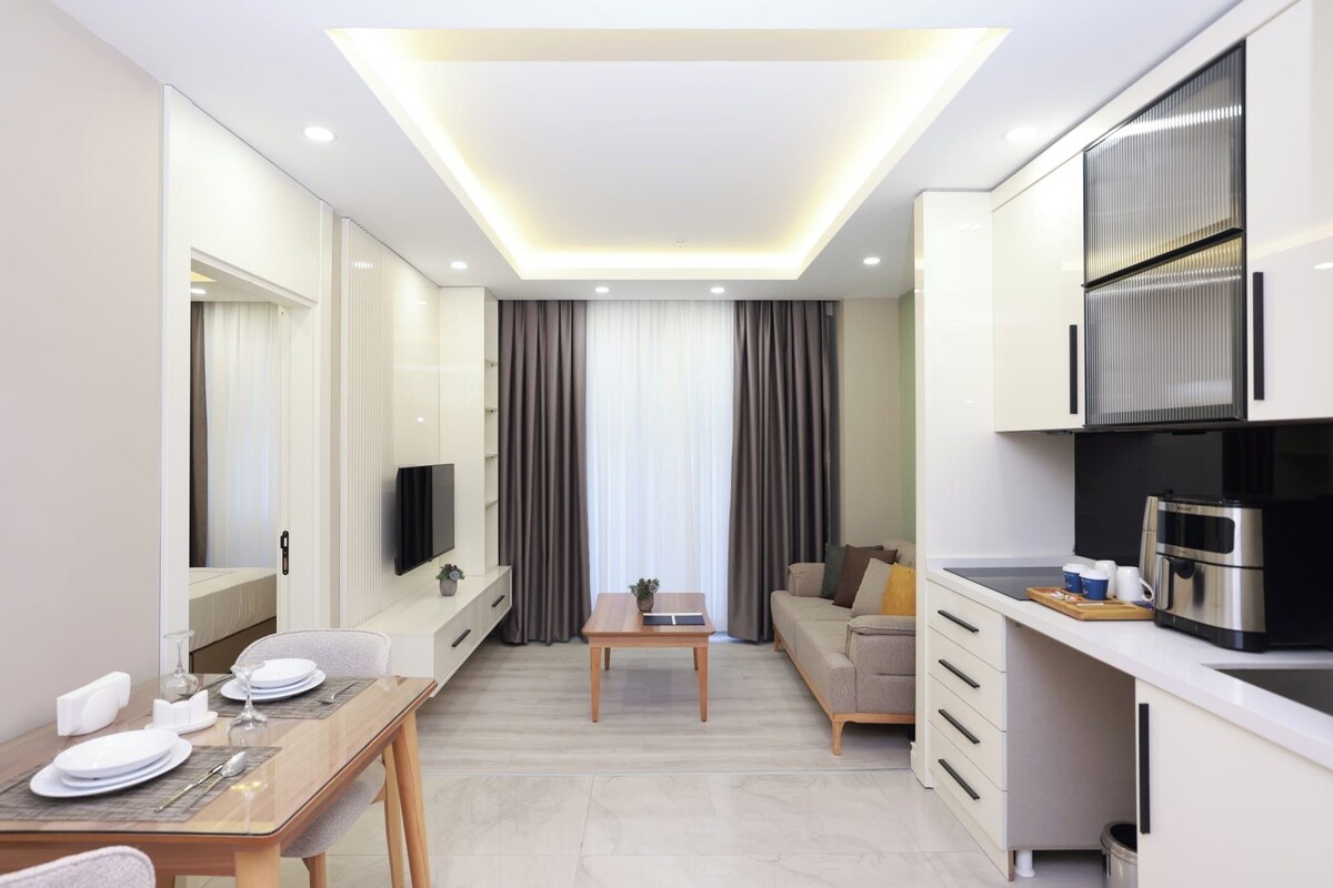 A modern hotel room with a dining area, kitchenette, and a large window with curtains.