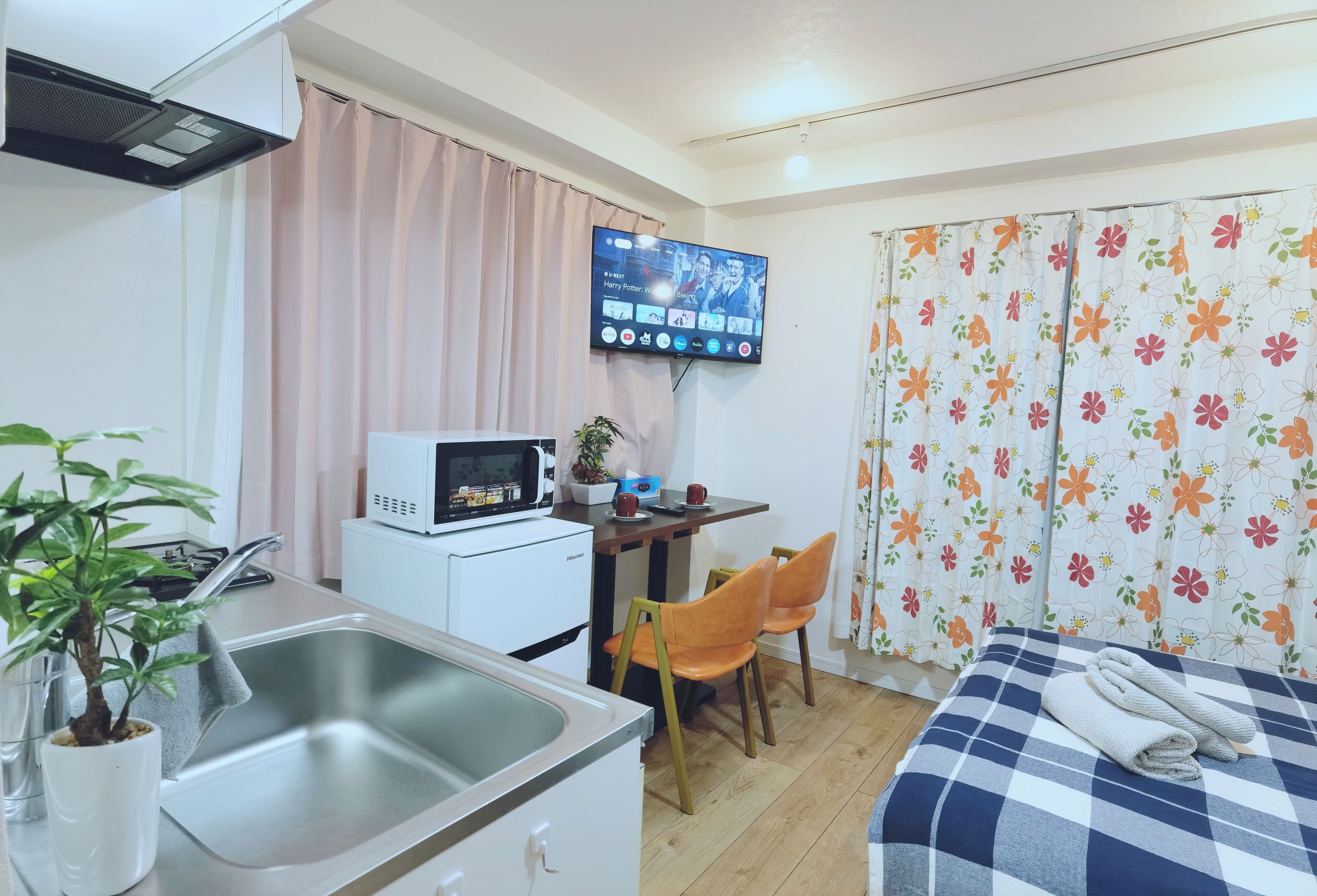 Une chambre d’hôtel compacte comprenant un coin cuisine, un lit recouvert d’une courtepointe à carreaux, un téléviseur fixé au mur et une fenêtre agrémentée de rideaux à motifs floraux.