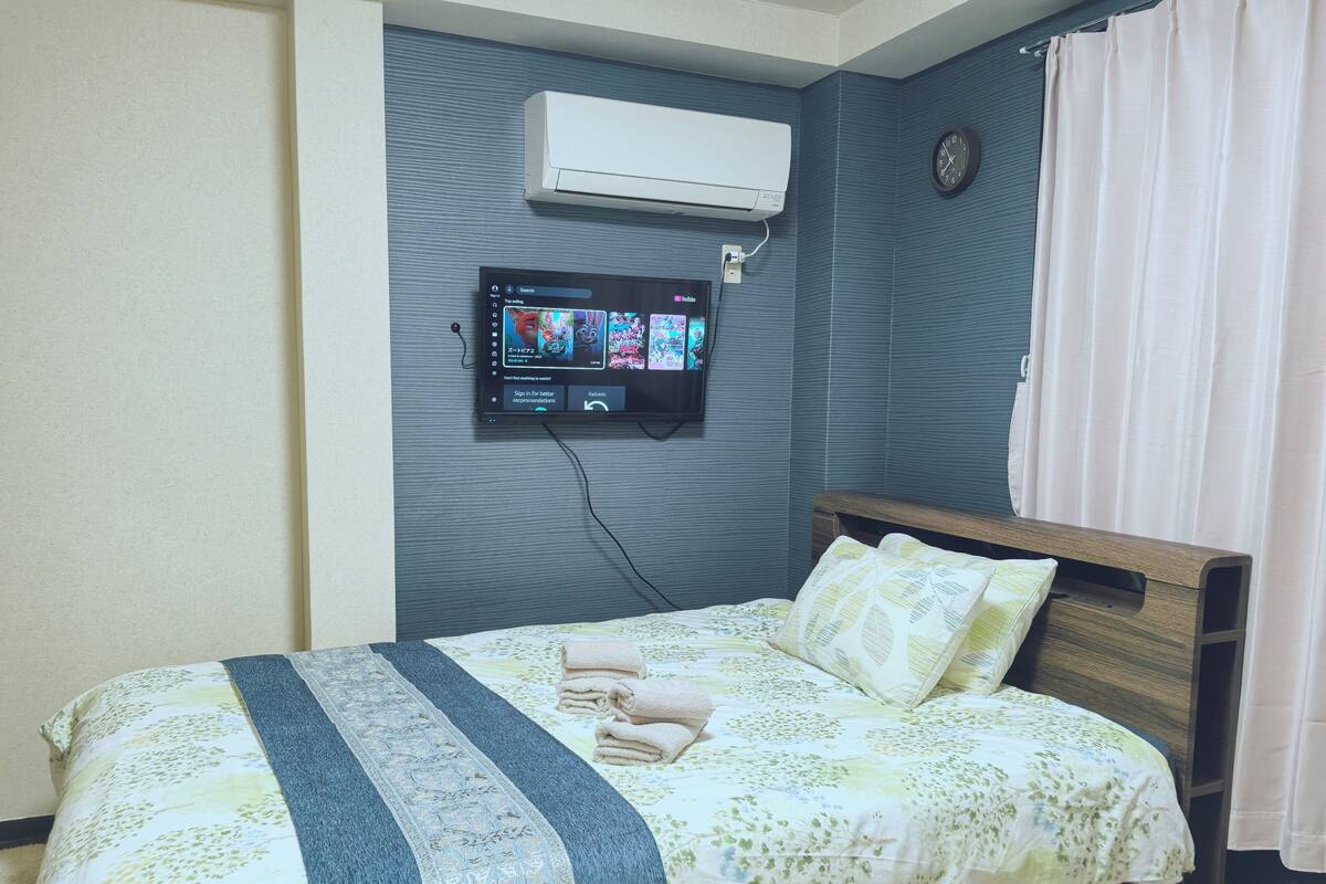 Une chambre d’hôtel équipée d’un lit, d’un téléviseur fixé au mur diffusant une vidéo, d’un climatiseur et d’une horloge murale.