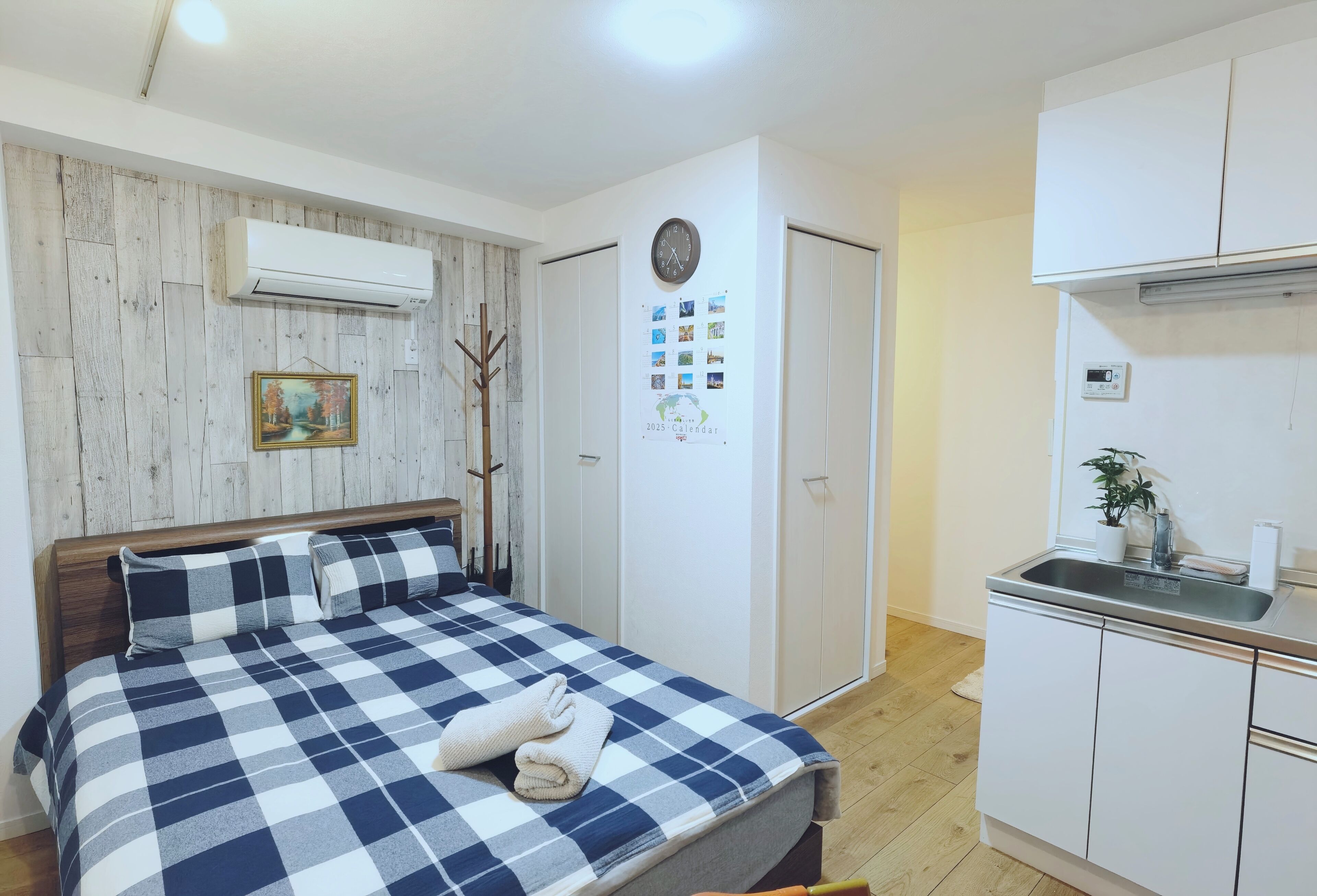 A bedroom with a checkered bedspread, a wooden headboard, an air conditioner, a wall-mounted clock, a calendar, and a small kitchenette with a sink and a potted plant.