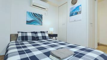 A neatly made bed with a checkered bedspread, a wall-mounted air conditioner, a painting, a clock, and a calendar.