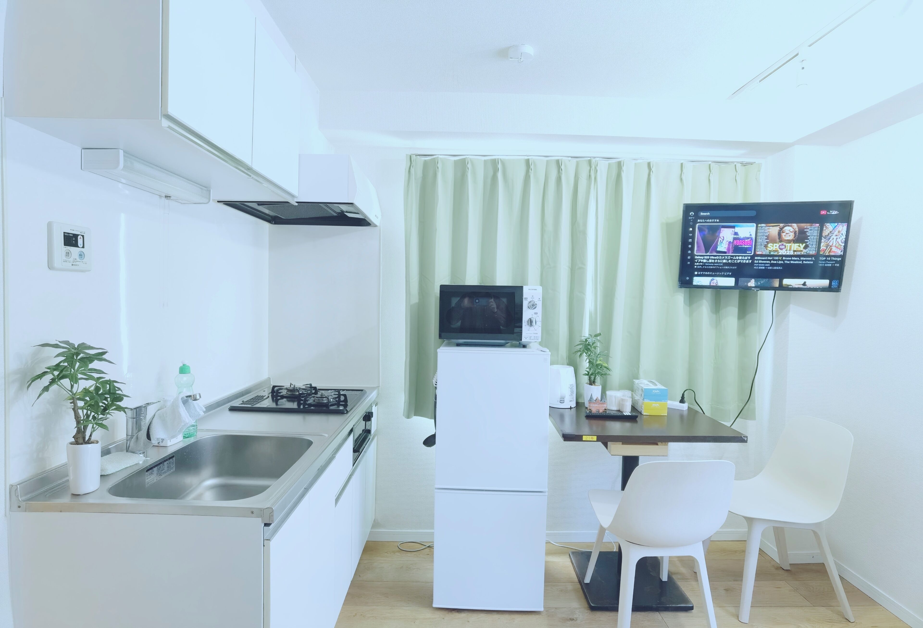 A compact kitchen with a gas stove, a refrigerator, a sink, and a flat-screen TV mounted on the wall.