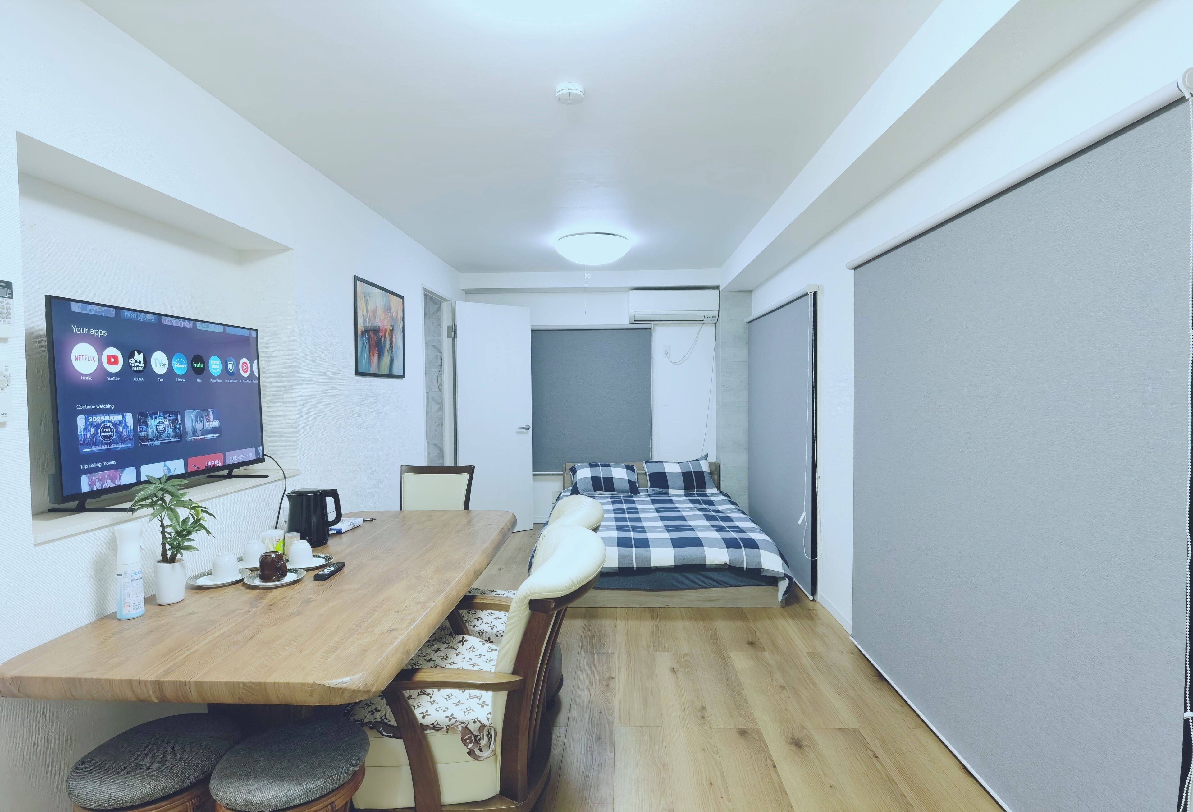 A compact room with a wooden dining table, a bed with a checkered blanket, a mounted TV displaying app icons, and a potted plant.
