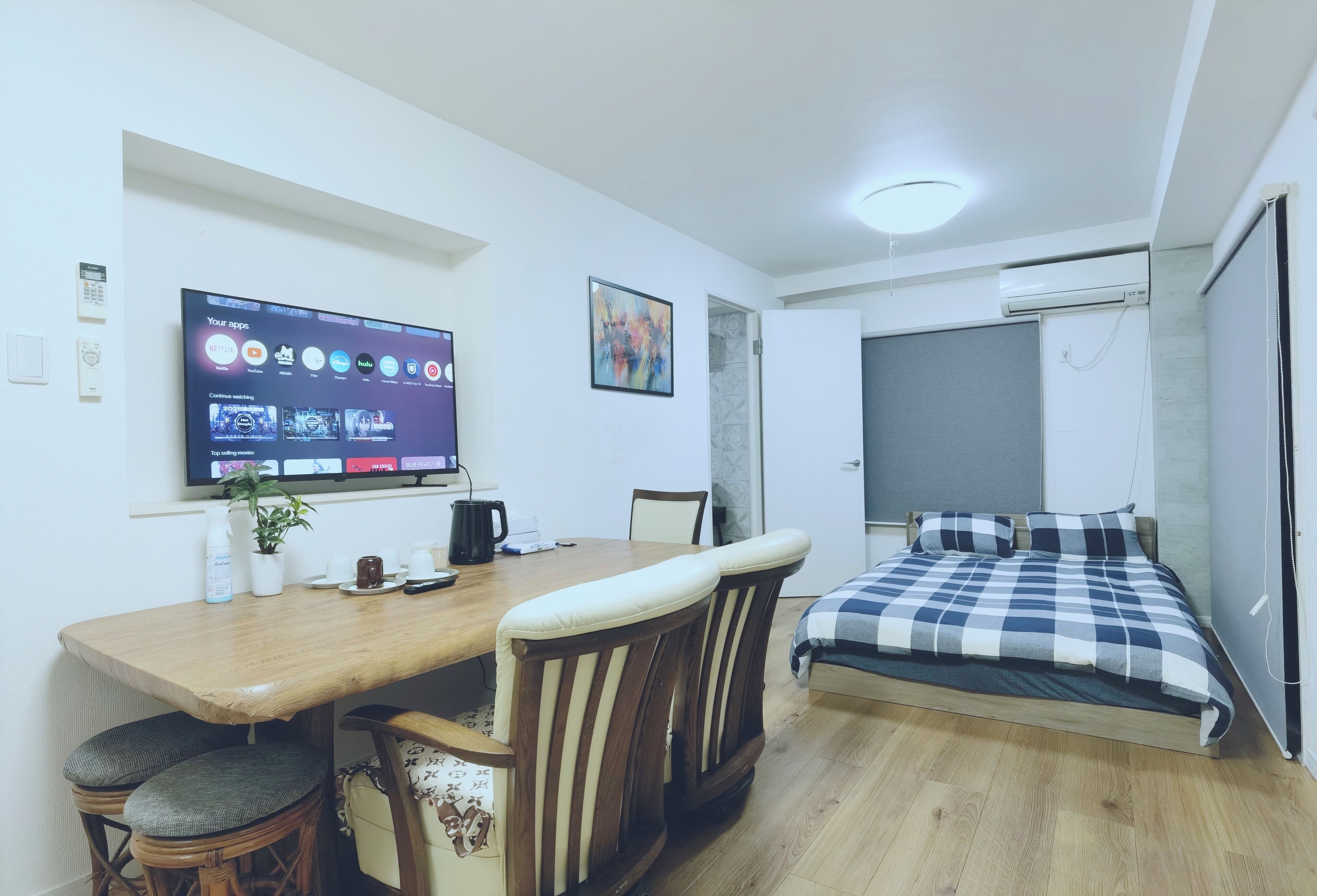 A compact room with a bed, a dining table with chairs, a TV mounted on the wall, and a small plant.