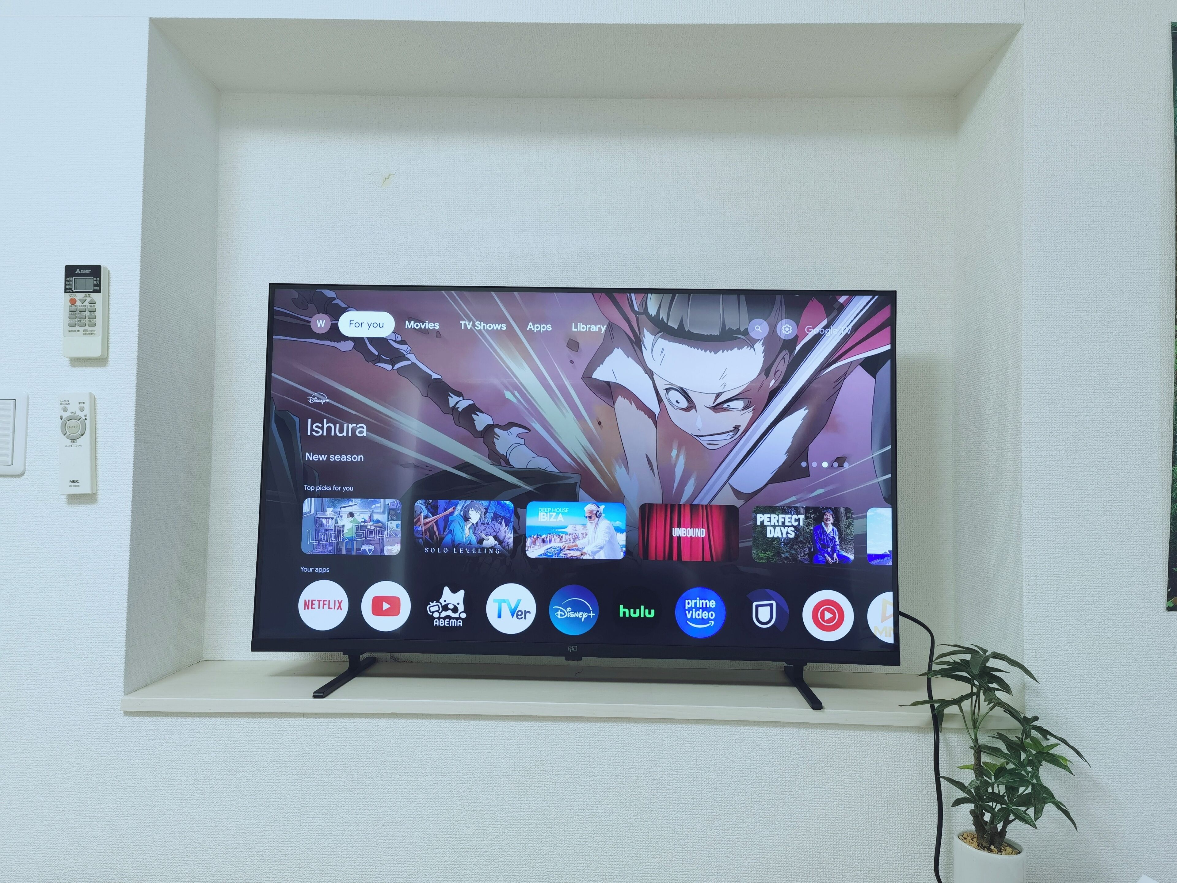 A television mounted on a wall with a remote control and a plant beside it.