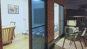 Ein Zimmer mit einer Schiebeglastür, die auf einen Balkon führt, einem Bett mit einem gestreiften Kopfkissen, einem Sessel und einer Klimaanlage.