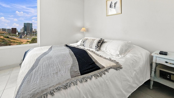 A bedroom with a bed, a nightstand, a lamp, and a city view.