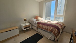 Ein Schlafzimmer mit einem Bett, einem Nachttisch mit Lampe, einem Fenster mit Blick auf Gebäude und einem schwarzen Teppich auf dem Boden.