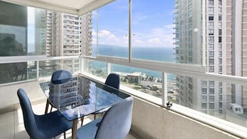 Un balcón con mesa y sillas de vidrio, con vista a un paisaje urbano y al mar.