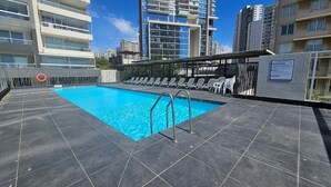 Piscina en la azotea con agua azul cristalina, rodeada de sillas de descanso y vistas a rascacielos.