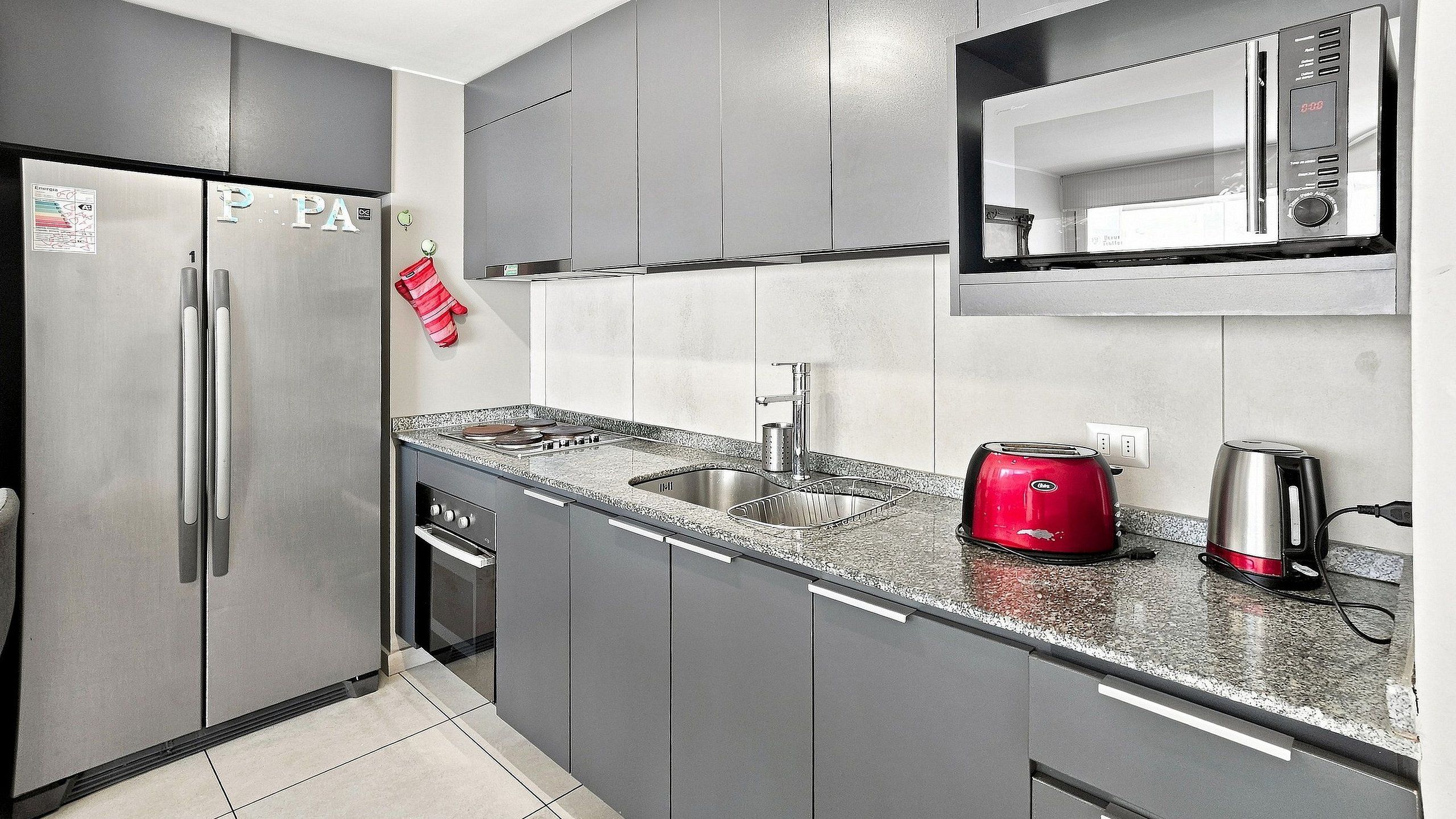 Una cocina con gabinetes grises, un refrigerador de acero inoxidable, una cafetera roja y un hervidor.