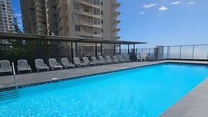 Una piscina con sillas de descanso y una zona cubierta, rodeada de edificios altos.