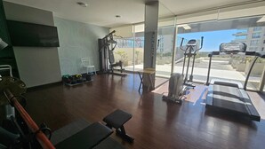 Un gimnasio con equipo de ejercicio, una banca y vistas a edificios a través de amplios ventanales.