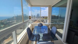 Ein Balkon mit Glastisch und Stühlen, der Blick auf eine Stadtlandschaft und den Ozean gewährt.