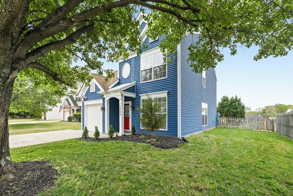 A two-story house with a garage, a red door, and a small front yard with a tree and shrubs.