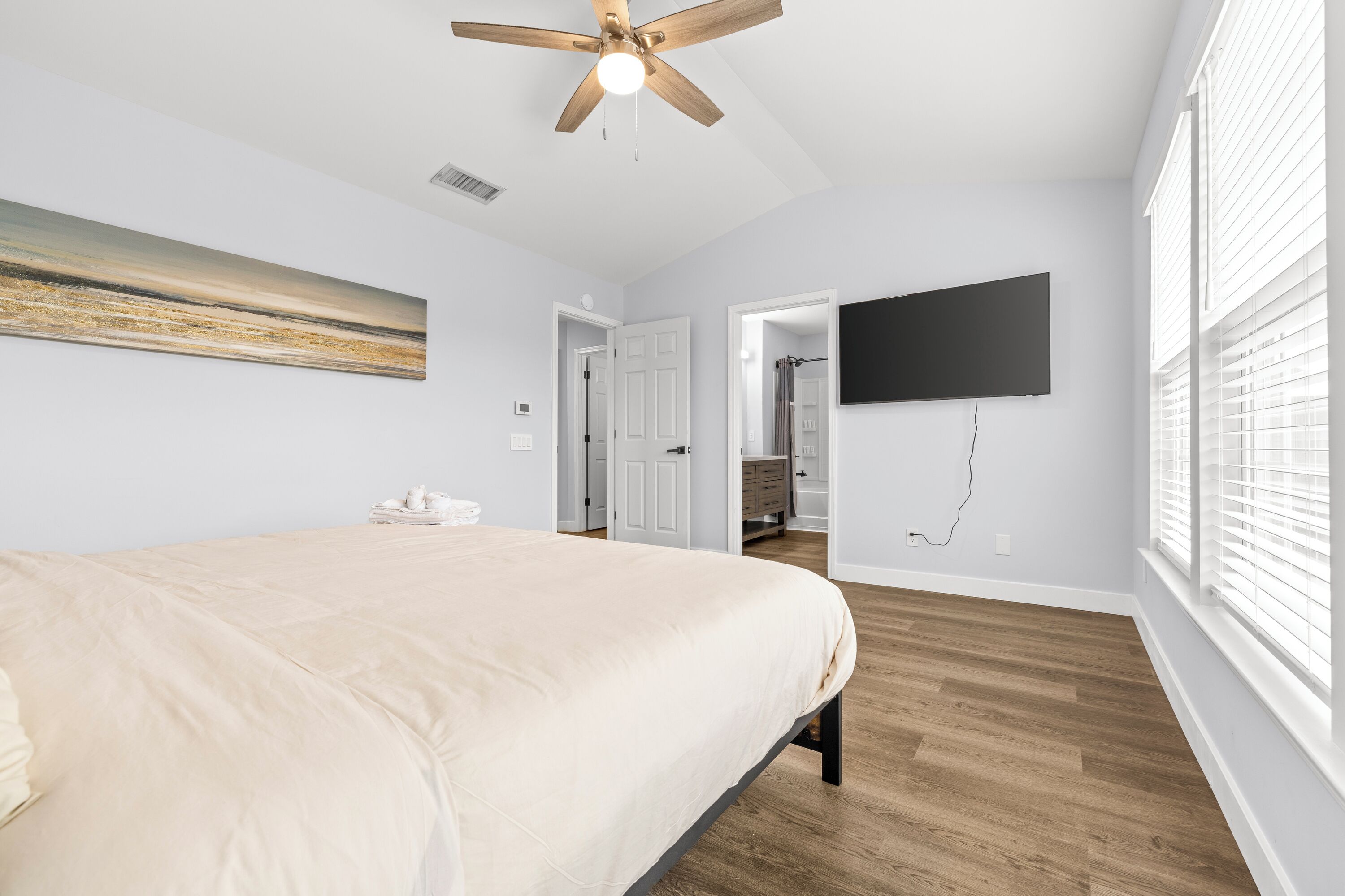 Un dormitorio con una cama grande, un ventilador de techo, un televisor de pantalla plana montado en la pared y una ventana con persianas.