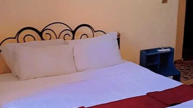 A bed with a white and red bedspread, a black metal headboard, and a blue chair in a room with a patterned carpet. - Clinton Hotel (Kikuyu)